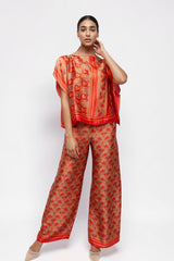Model wearing ALBANA New York silk wide leg pants and matching top in vibrant orange and red floral scroll print.