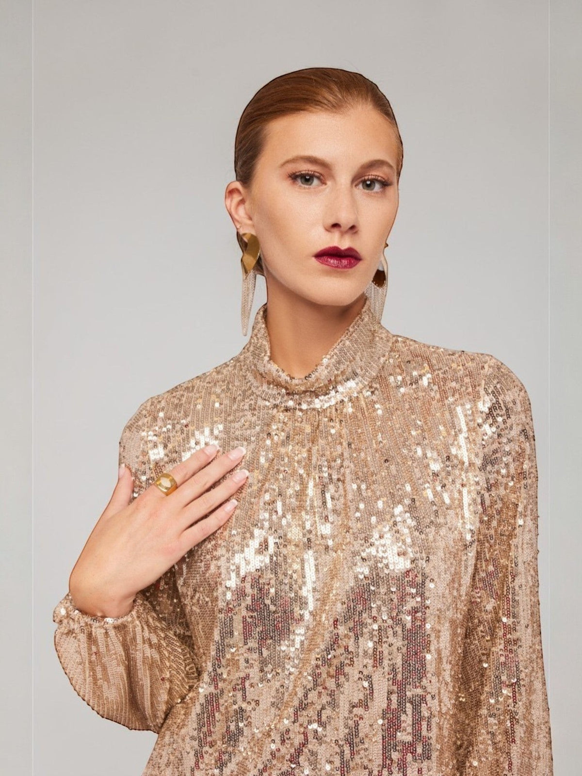 ALBANA New York gold sequin long sleeve blouse with a mock neck worn by a model with gold statement earrings